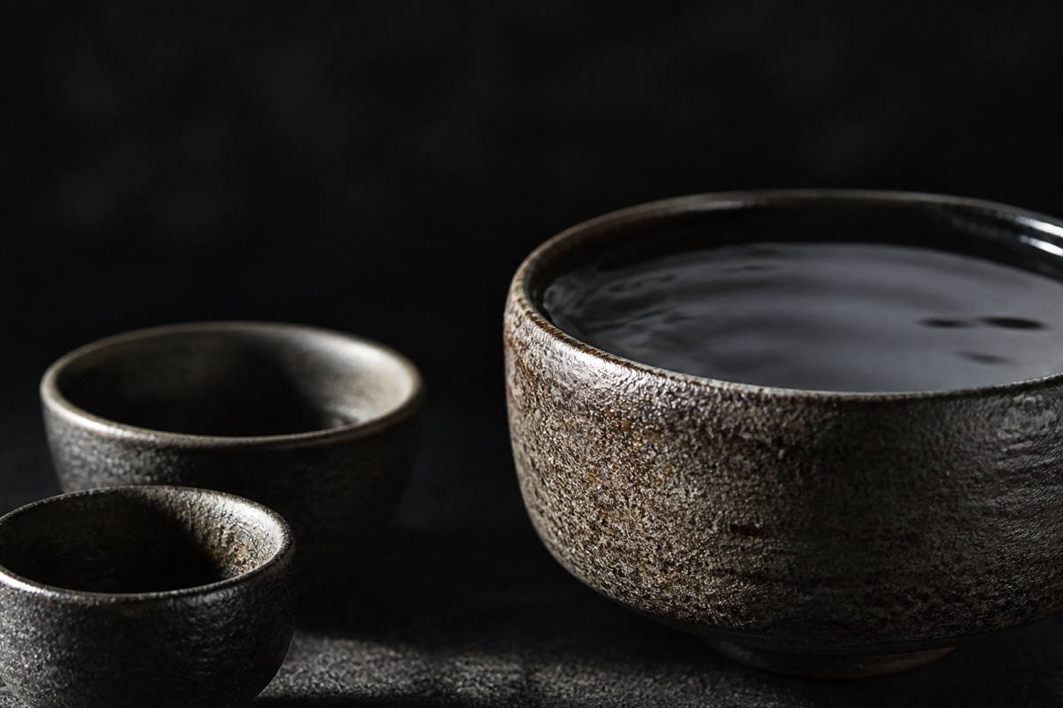 어두운 배경의 극적 명암 정물 — 크기가 다른 세 개의 그릇이 나란히 놓여 있고 가장 큰 그릇에만 물이 가득 담겨 빛을 받는 매크로 — 같은 물줄기라도 그릇의 크기만큼 담긴다는 은유, 글자 없음.